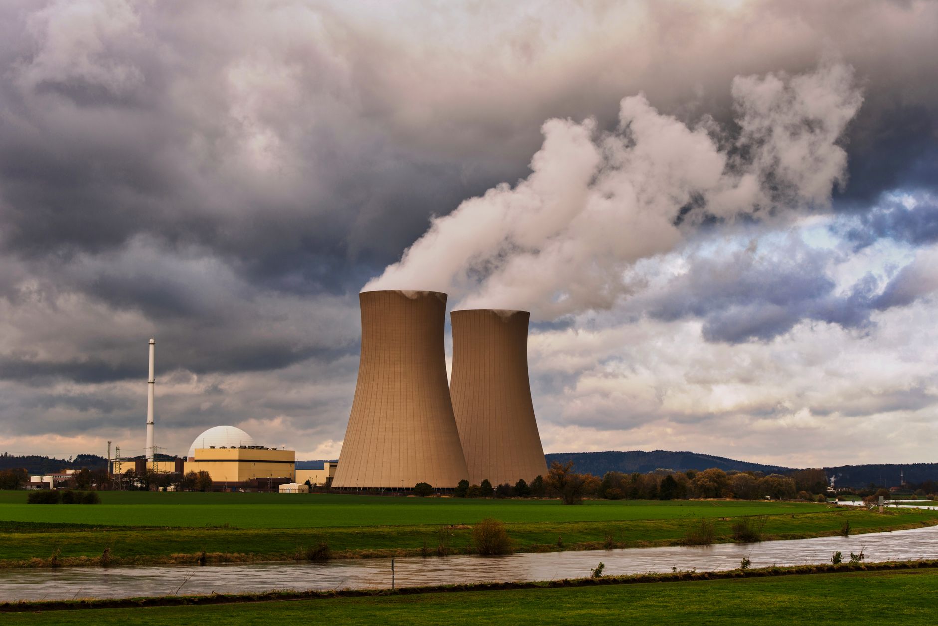 Centrale nucléaire avec deux tours de refroidissement qui émettent de la vapeur, au-dessus d’une rivière et de prairies, sous un ciel nuageux.