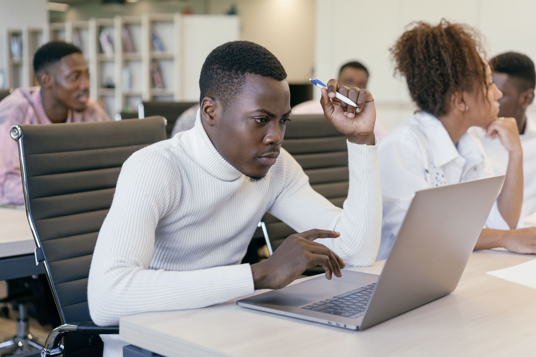 Un jeune homme en pull blanc travaille sur un ordinateur portable, un stylo à la main, dans un espace de bureau lumineux avec d’autres personnes.